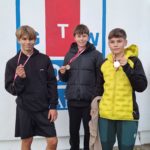 Trzech chłopców pozuje z medalami przed ścianą z logo klubu wioślarskiego. Uśmiechnięci, ubrani sportowo, dumni z osiągnięć.