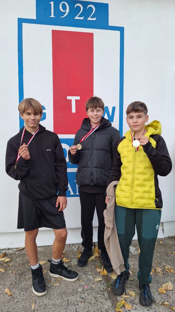 Trzech chłopców pozuje z medalami przed ścianą z logo klubu wioślarskiego. Uśmiechnięci, ubrani sportowo, dumni z osiągnięć.