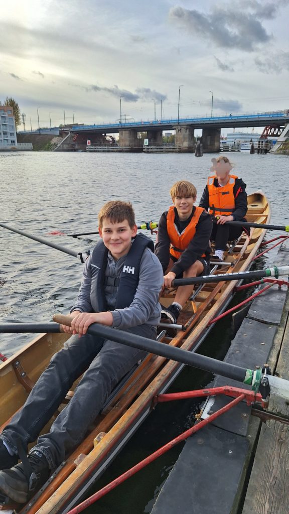 Trzech chłopców w kamizelkach asekuracyjnych siedzi w drewnianej łodzi wiosłowej przy pomoście, gotowi do treningu na wodzie. W tle most i zabudowania nad rzeką.