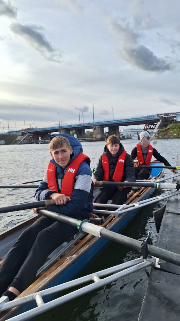 Trzech chłopców siedzi w łodzi wiosłowej na wodzie, w kamizelkach asekuracyjnych, gotowa do wspólnego wiosłowania. W tle most i rzeka.