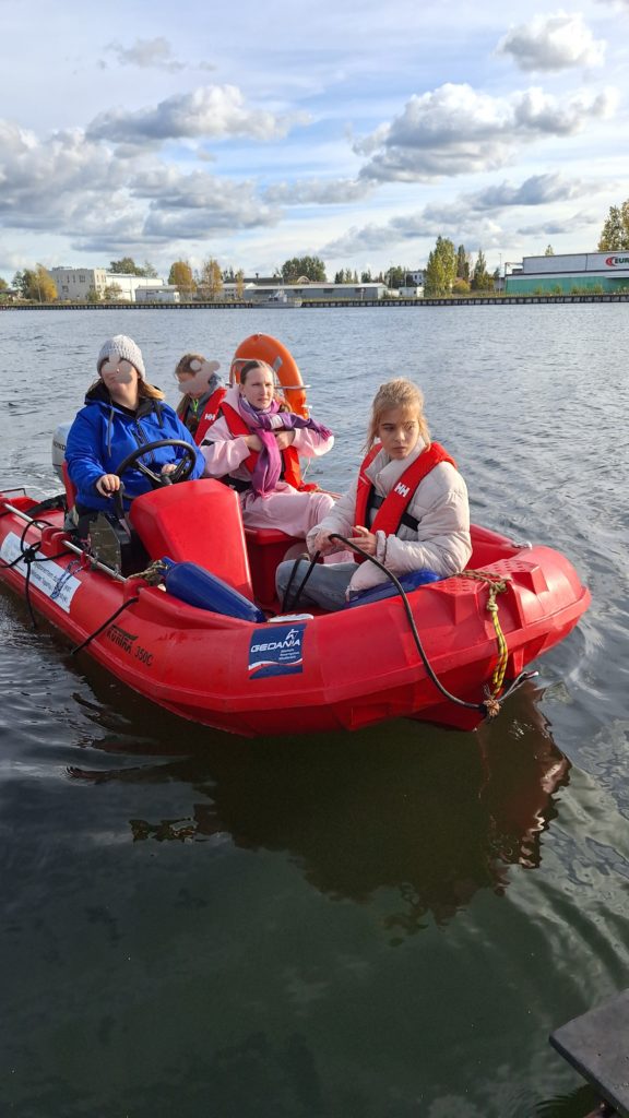 Cztery dziewczyny w kamizelkach ratunkowych siedzi w małej motorówce na spokojnej wodzie. Jedna z dziewczyn trzyma kierownicę.