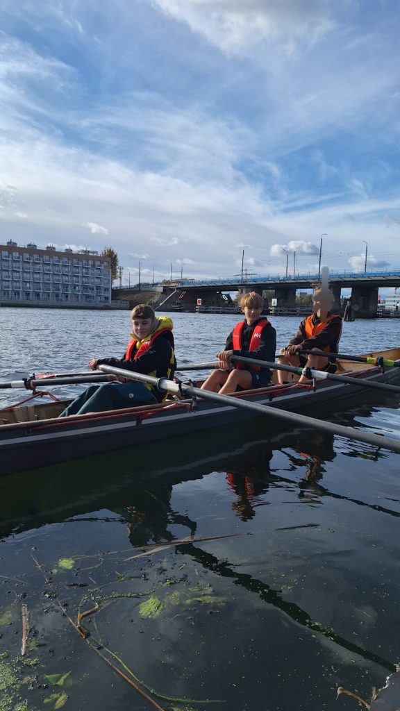 Trzech chłopców siedzi w łodzi wiosłowej na wodzie, w kamizelkach asekuracyjnych, gotowa do wspólnego wiosłowania. W tle most i rzeka.