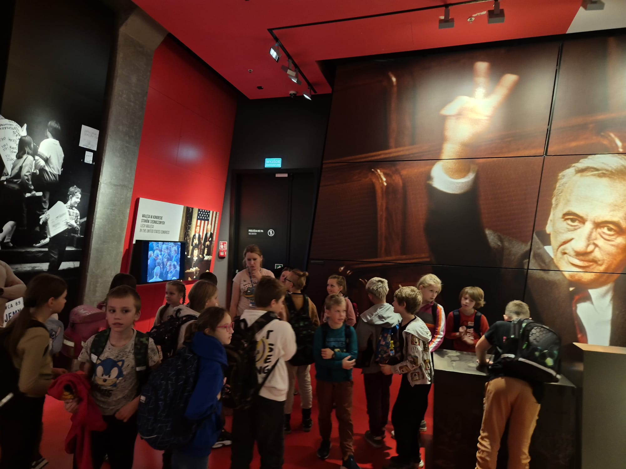 W sali wystawowej stoi szesnaście dzieci. Dzieci słuchają co mówi do nich Pani edukatorka .Naprzeciwko dzieci znajduje się zdjęcie Tadeusza Mazowieckiego. Po prawej stronie s jest monitor i tablica ze zdjęciem polityków.