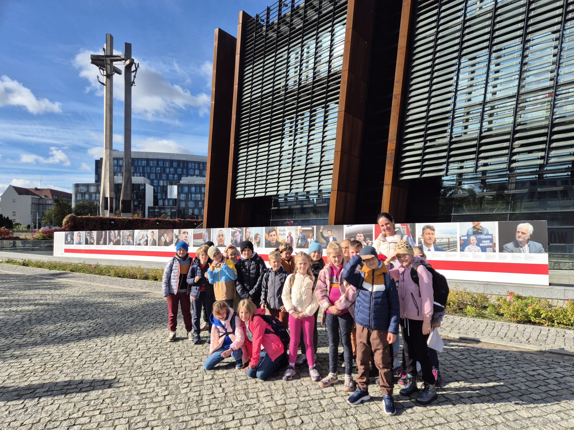 Dziewiętnaście dzieci stoi obok siebie Są ubrane jesiennie w czapki i kurtki. Za dziećmi znajduje się Europejskie Centrum Solidarności. Obok budynku widać trzy krzyże.