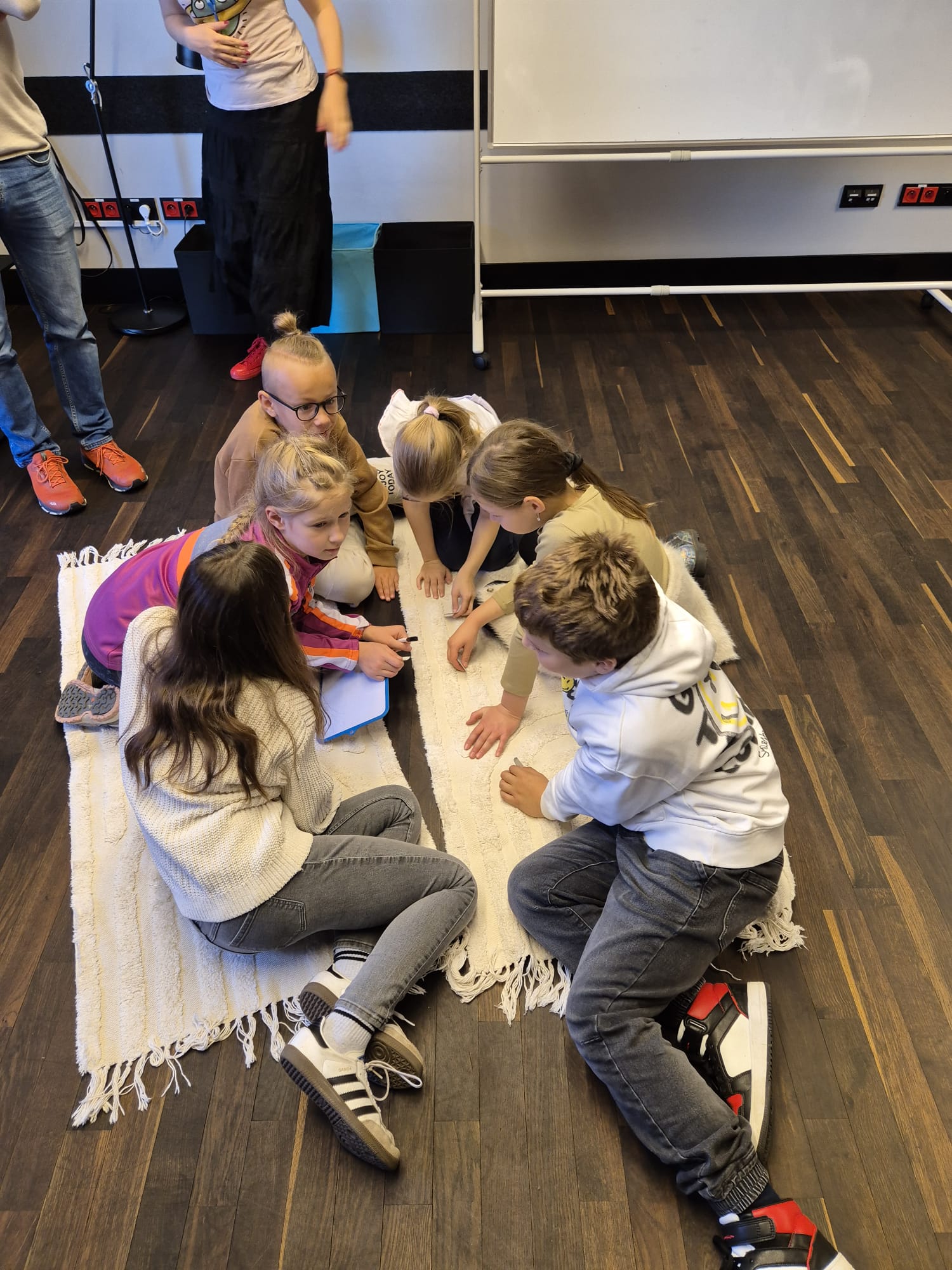 na dywanikach siedzą dzieci. Jest ich siedem. Dzieci wymyślają nazwę dla swojej grupy Jedna dziewczynka ma w ręce notatnik i długopis, aby zapisać nazwę.