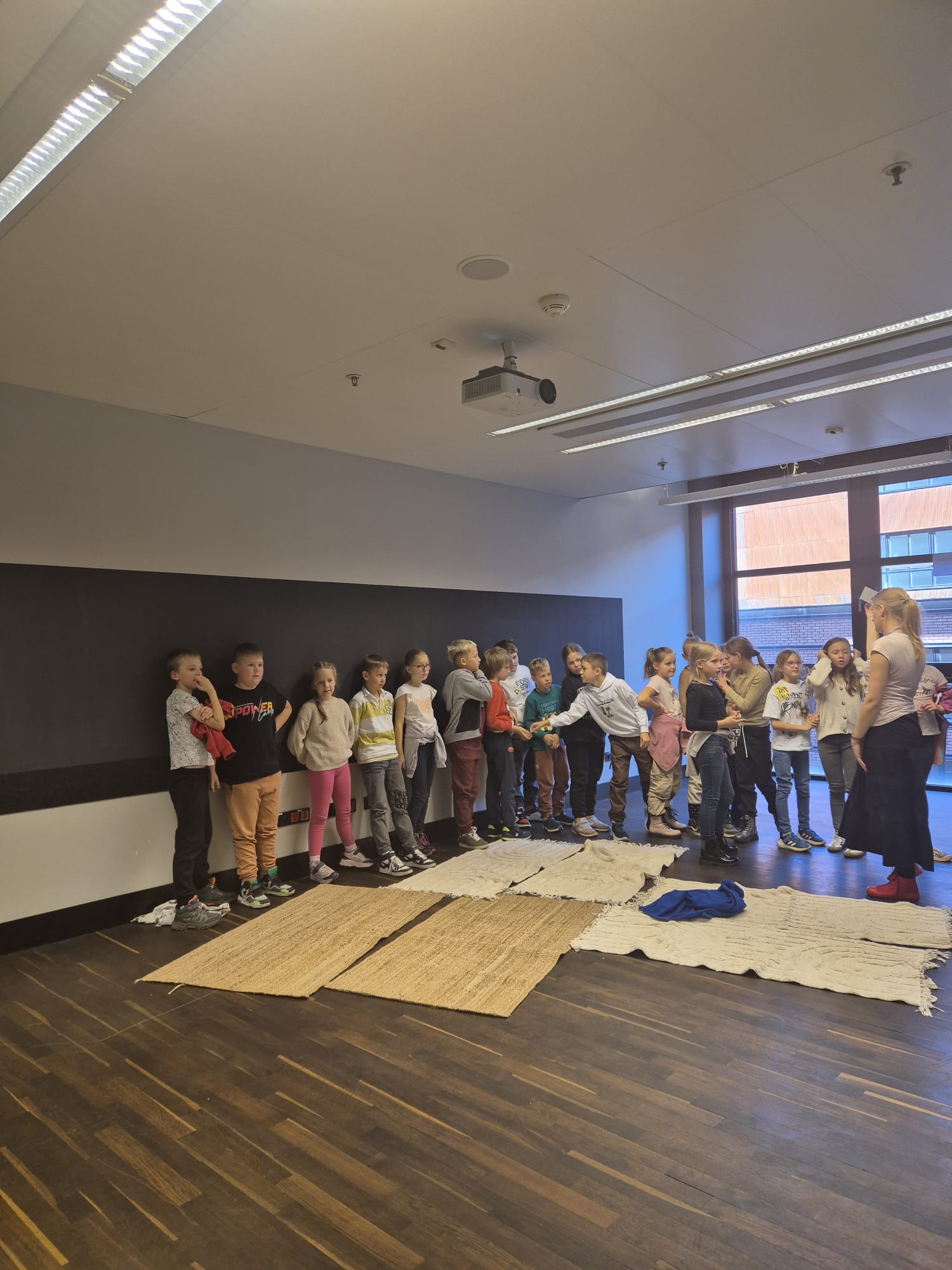 Dzieci stoją jedno za drugim. jest ich dziewiętnaście. Będą głosowały i oddawały głos na którą nazwę grupy się zdecydują. Obok dzieci stoi Pani w długiej spódnicy. Która tłumaczy dzieciom zasady głosowania.