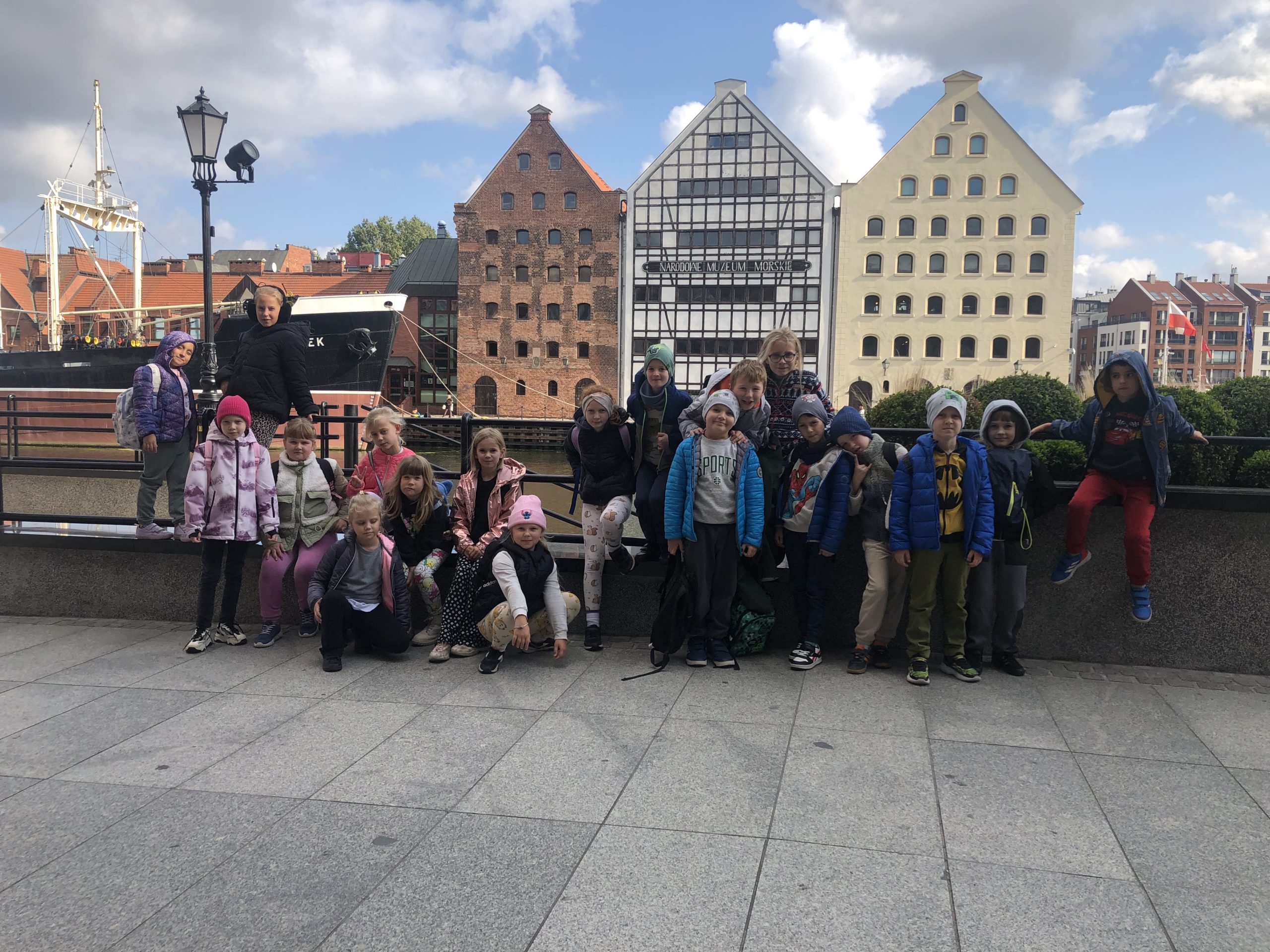Grupa 20 uczniów stoi na nabrzeżu, w tle znajduje się marina oraz budynek Muzeum.