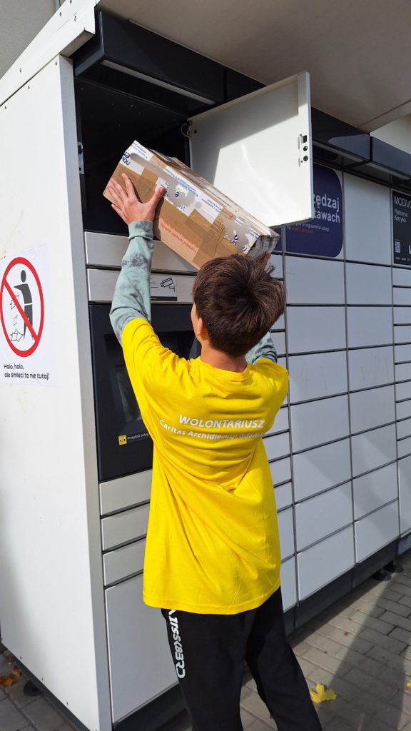 Chłopiec w koszulce wolontariusza wkłada kartonowe pudełko do górnej skrytki paczkomatu.