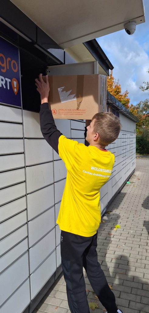 Chłopiec w koszulce wolontariusza umieszcza duże pudło w górnej komorze paczkomatu na tle jesiennych drzew.