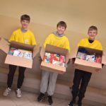 Trzech uśmiechniętych chłopców w koszulkach wolontariusza stoi w pomieszczeniu i trzyma otwarte pudła wypełnione przyborami szkolnymi.