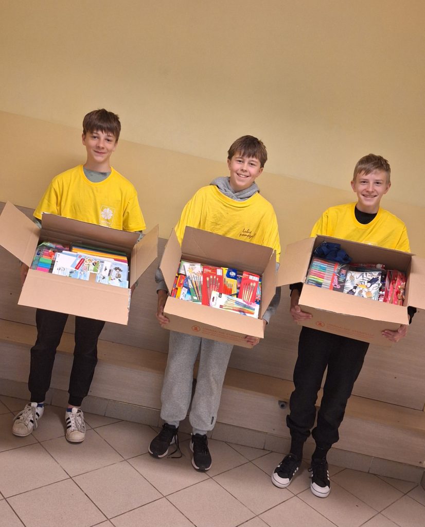 Trzech uśmiechniętych chłopców w koszulkach wolontariusza stoi w pomieszczeniu i trzyma otwarte pudła wypełnione przyborami szkolnymi.