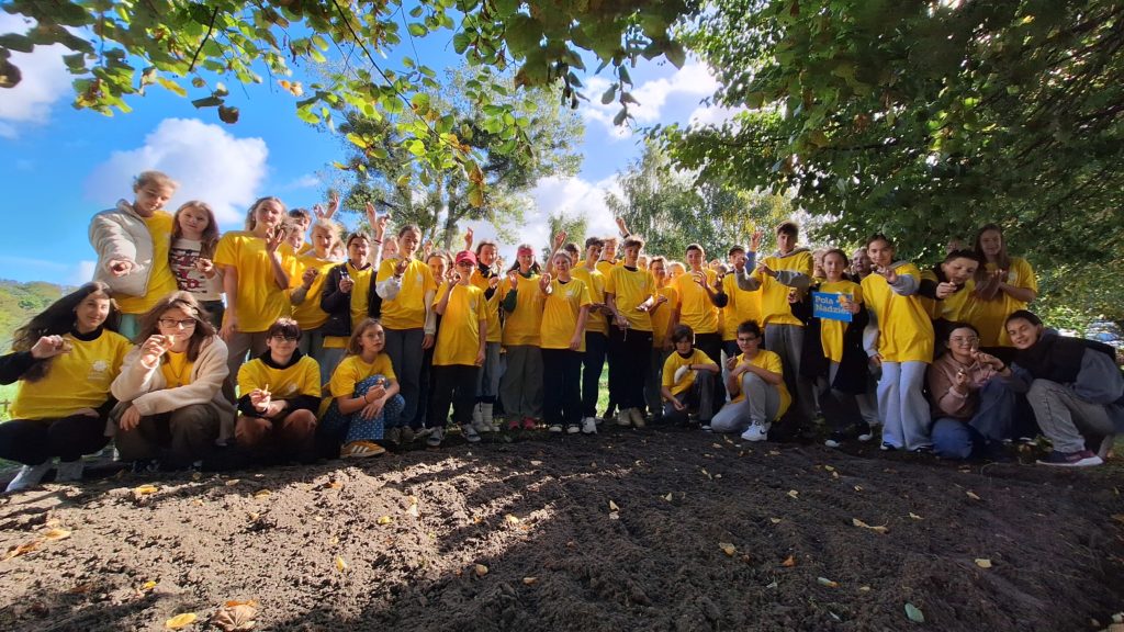 Duża grupa młodych osób ubranych w koszulki wolontariuszy, stojących na świeżym powietrzu pod drzewami. Wszyscy pozują do wspólnego zdjęcia, uśmiechają się i trzymają w dłoniach cebulki kwiatów. Na środku jest tabliczka z napisem „Pola Nadziei”.