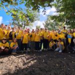 Duża grupa młodych osób ubranych w koszulki wolontariuszy, stojących na świeżym powietrzu pod drzewami. Wszyscy pozują do wspólnego zdjęcia, uśmiechają się i trzymają w dłoniach cebulki kwiatów. Na środku jest tabliczka z napisem „Pola Nadziei”.