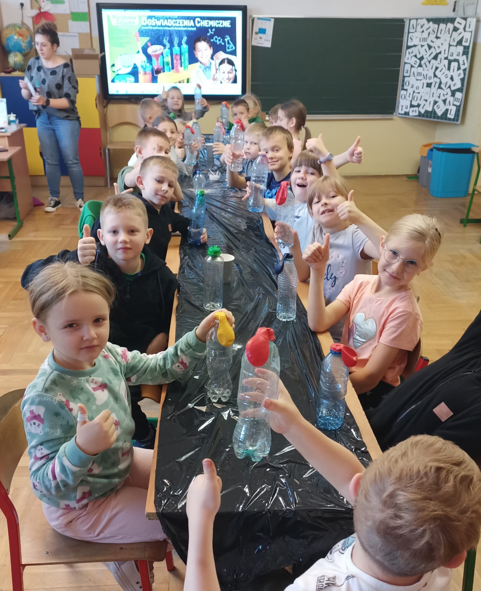 Klasa edukacji wczesnoszkolnej siedzi przy długim stole osłoniętym czarną folią. Dzieci trzymają plastikowe butelki z założonymi kolorowymi balonami i pokazują kciuki w górę; w tle nauczycielka i monitor z planszą „Doświadczenia chemiczne"