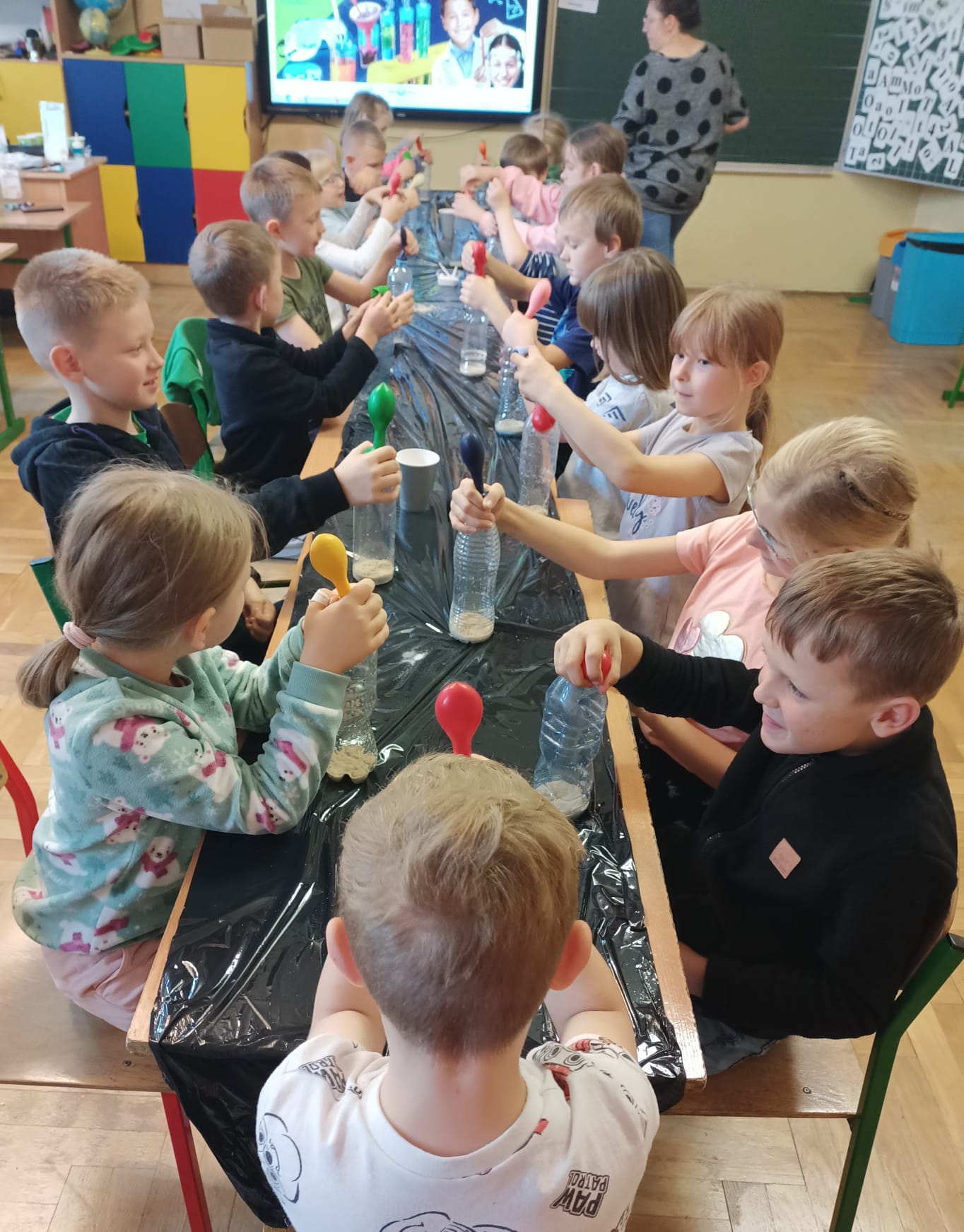 Dzieci siedzą w dwóch rzędach przy długim stole zabezpieczonym czarną folią i wykonują doświadczenie: na szyjkach plastikowych butelek mają założone kolorowe balony, które zaczynają się napełniać
