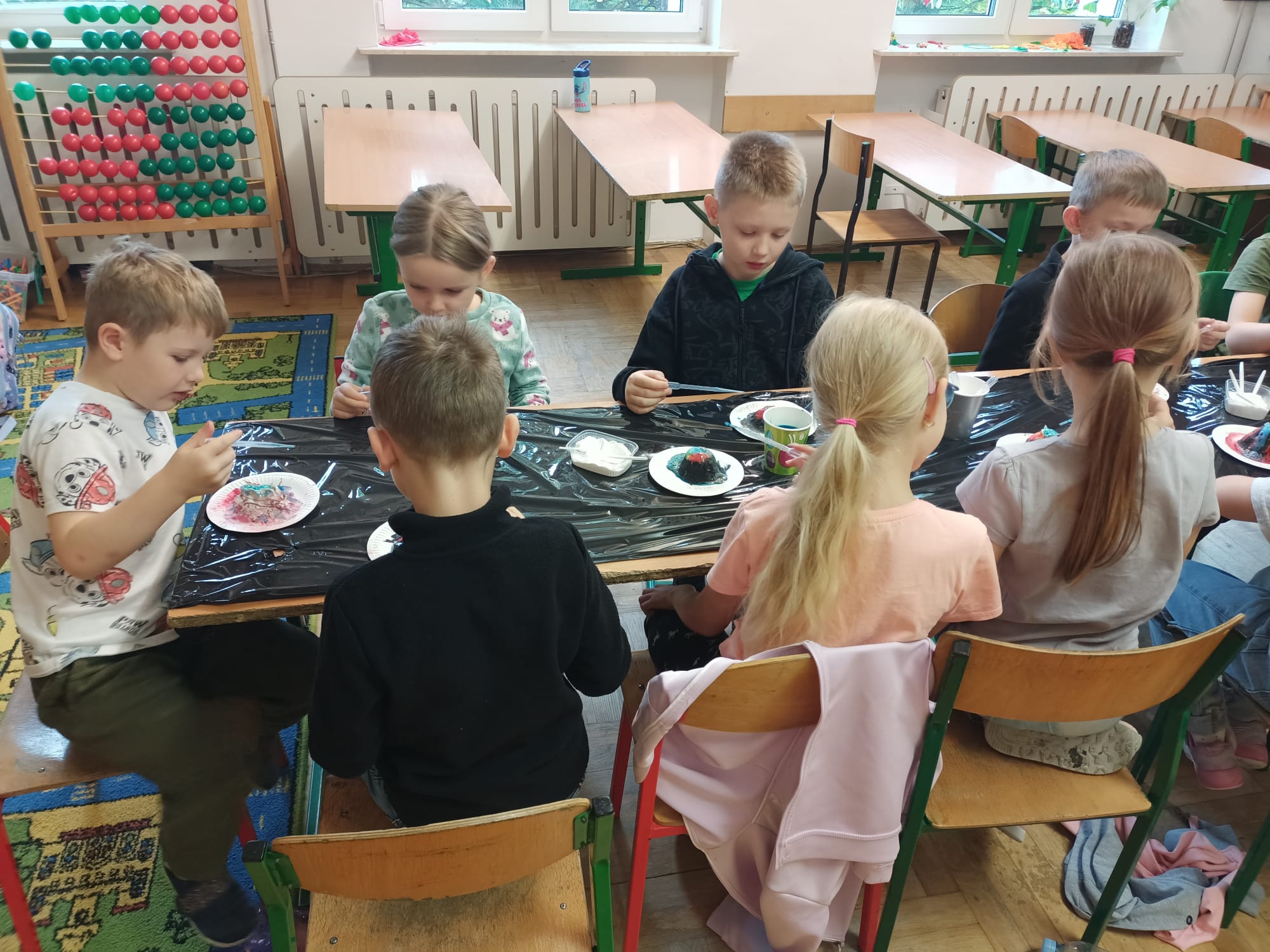 Grupa dzieci siedzi przy stole zabezpieczonym czarną folią i wykonuje doświadczenie na modelach miniwulkanow umieszczonych na papierowych talerzykach. Uczniowie dozują barwne roztwory minipipetami, obok mają kubeczki z materiałami; w tle widać okna, ławki i liczydło. 
