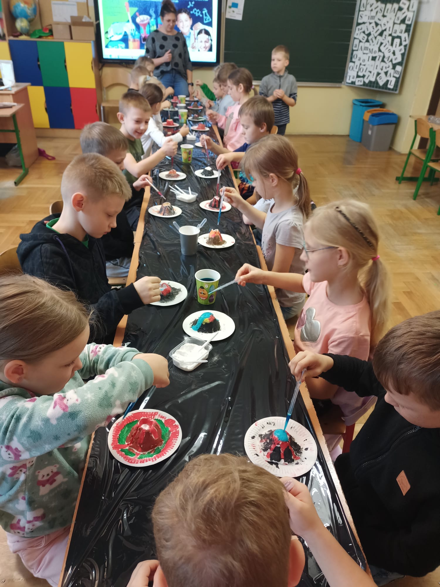 Uczniowie klas I–III pracują przy długim stole przykrytym folią. Na talerzykach mają modele wulkanów a za pomocą pipet Pasteura nanoszą kolorowe ciecze do wnętrza modeli. Z wnętrza wulkanów wydobywa się piana imitująca lawę. Na końcu sali znajduje się nauczyciel, który czuwa nad bezpieczeństwem doświadczeń. 
