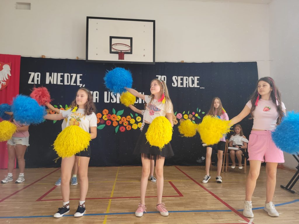 Grupa dziewcząt w sportowych strojach tańczy z żółto - niebieskimi pomponami.