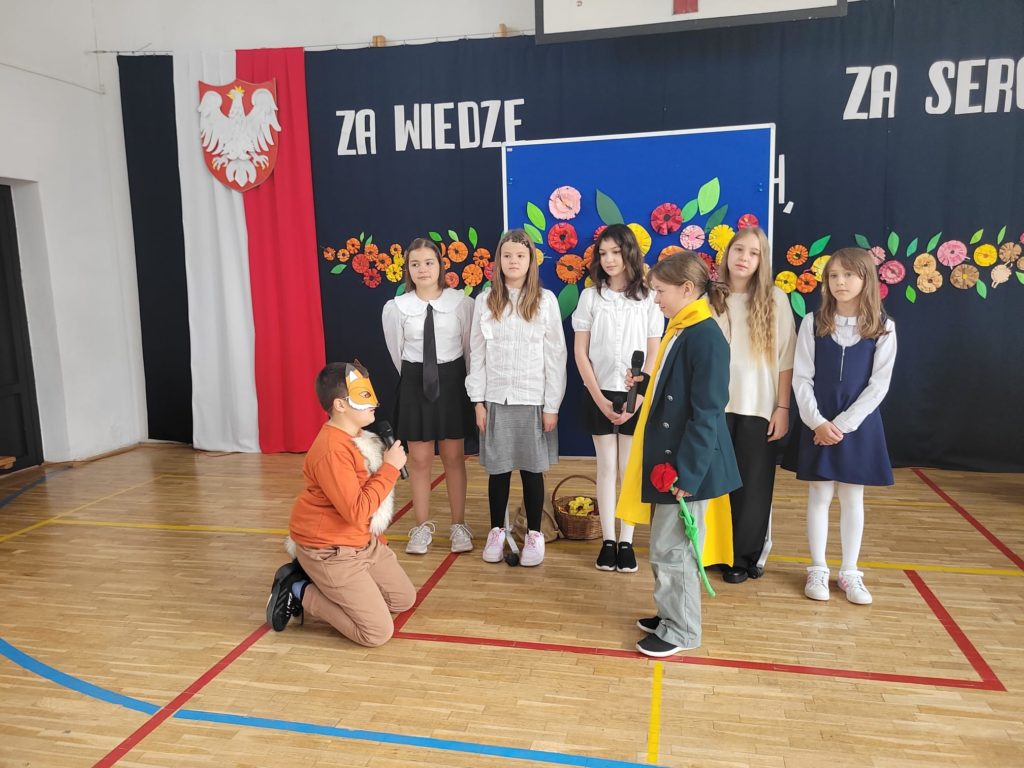 Dziewczynka przebrana za Małego Księcia z różą w dłoni stoi przodem do klęczącego chłopca, który jest przebrany za lisa. Z tyłu grupa dziewcząt, które stoją w szeregu.