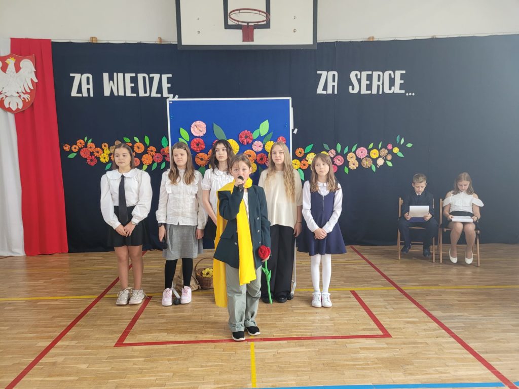 Grupa dziewcząt stoi na tle dekoracji z kwiatów oraz napisu "Za wiedzę, za uśmiech, za serce". Przed nimi dziewczynka przebrana za Małego Księcia.