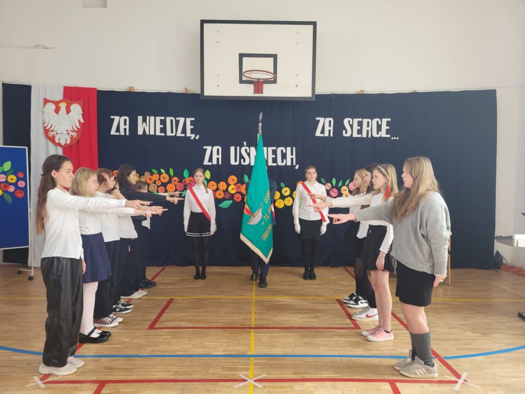 Dziewczęta i chłopcy stoją w dwóch kolumnach. Mają prawe ręce skierowane w stronę sztandaru, który jest pośrodku nich.