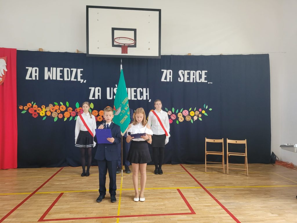 Dziewczynka i chłopiec stoją z mikrofonami w dłoniach. Za nimi poczet sztandarowy. 