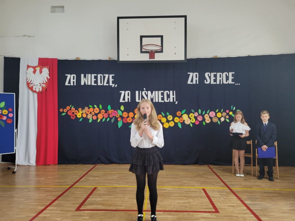 Dziewczynka ubrana w strój galowy stoi na tle dekoracji z kwiatów i mówi do mikrofonu.