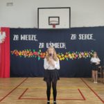 Dziewczynka ubrana w strój galowy stoi na tle dekoracji z kwiatów i mówi do mikrofonu.