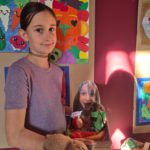 Uczennica stoi przy swojej pracy plastycznej przedstawiającej kolorowe owoce i warzywa. W dłoniach trzyma kartonowe pudełko z pracą konkursową. W tle widać inne prace plastyczne o tematyce owoców i warzyw.