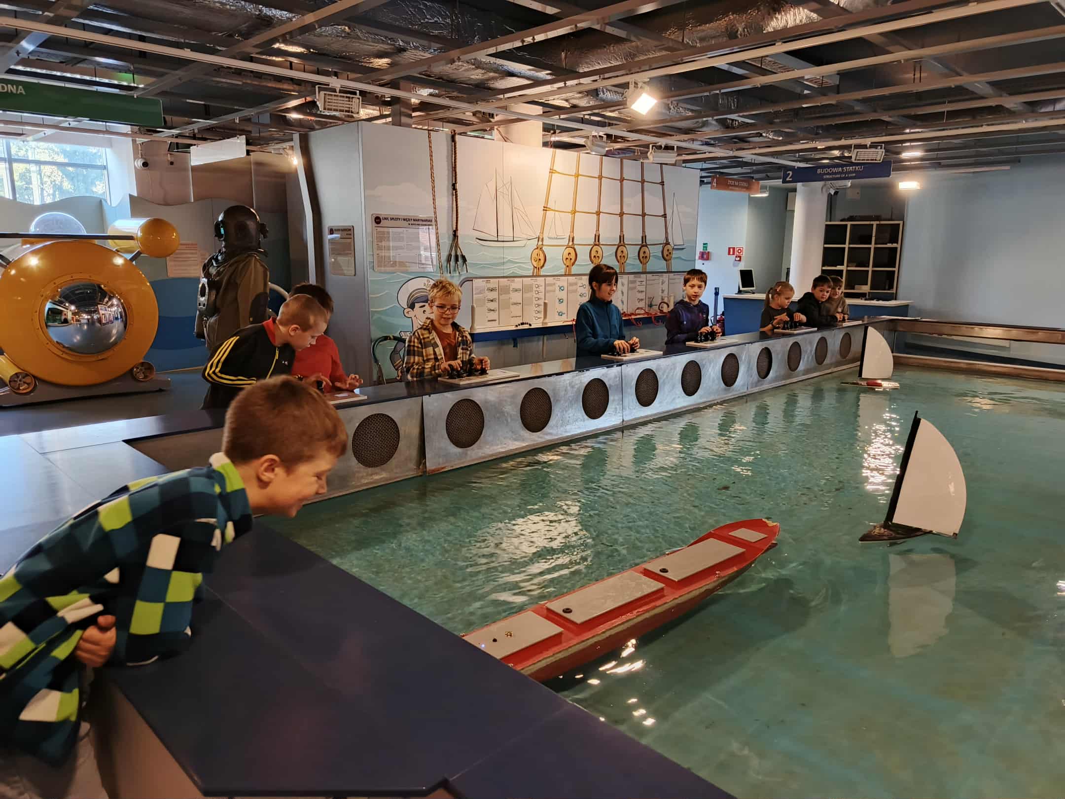 Kilku chłopców stoi przy dużym basenie z wodą. Każdy z nich trzyma pilota i steruje małym modelem statku lub żaglówki. Modele pływają po wodzie — widać jeden większy statek z czerwonym pokładem i dwie małe żaglówki. Na ścianie za dziećmi znajdują się rysunki i plansze z obrazkami statków.