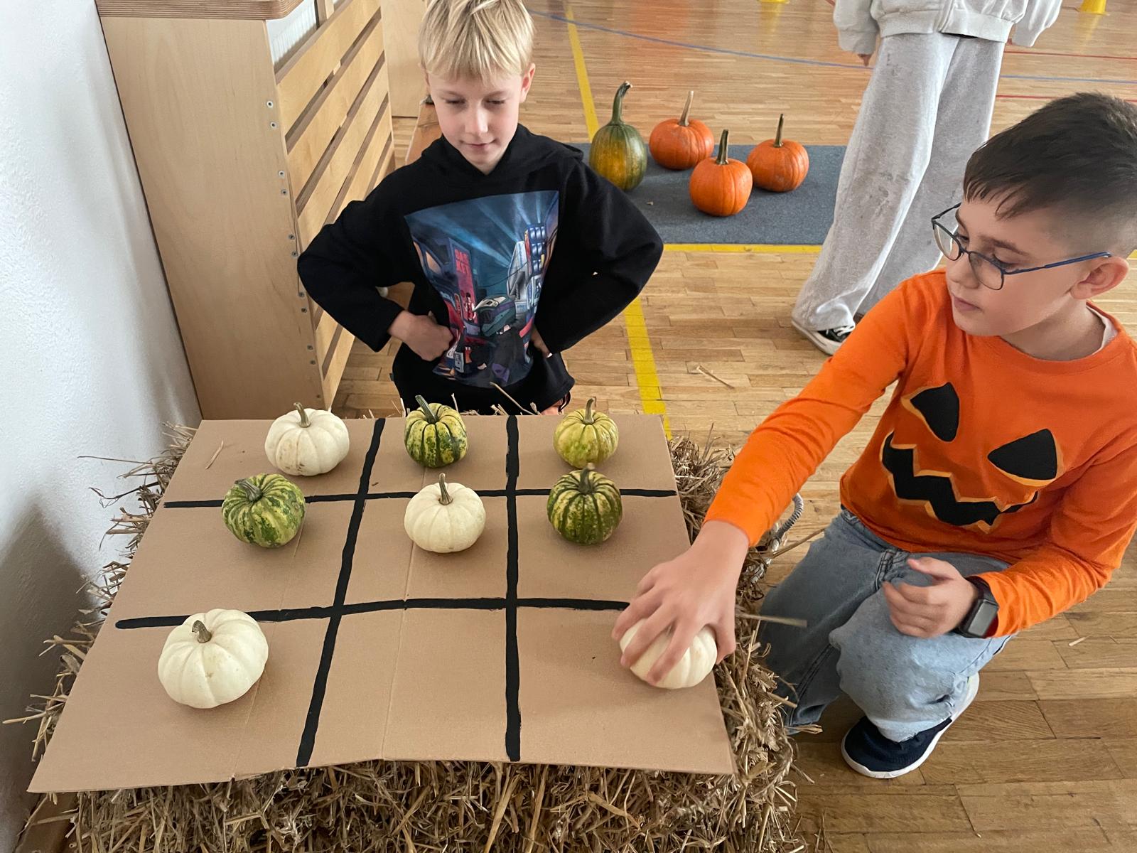  Dwaj chłopcy grają w "Kółko i Krzyżyk" z użyciem małych ozdobnych dyń i tykw. Plansza jest narysowana czarną farbą na kawałku tektury położonym na balocie siana. Chłopiec w pomarańczowej koszulce z motywem dyni kładzie jeden z jasnych pionków.