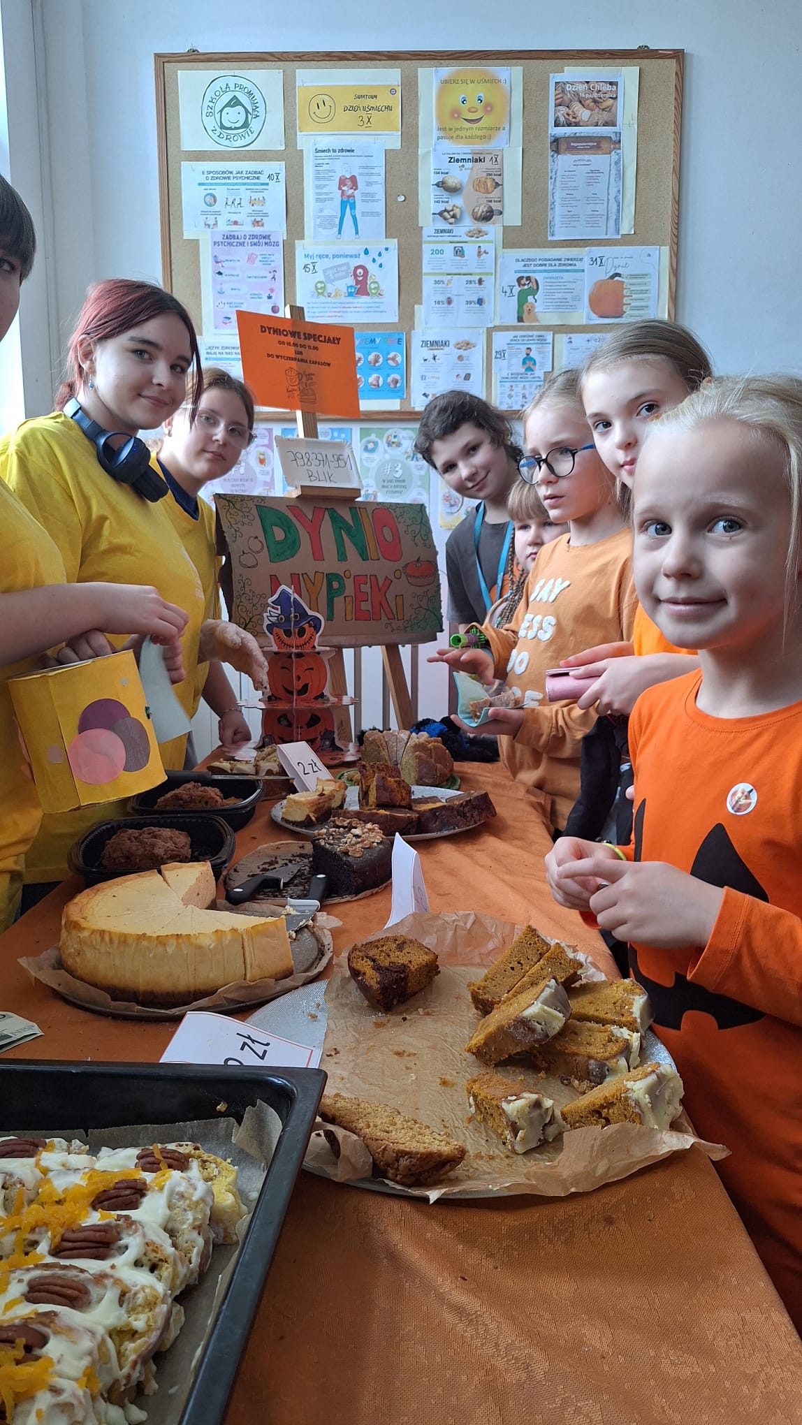 Kilka dzieci i nastolatków stoi za stołem z ciastami i wypiekami. Na pierwszym planie widać różne rodzaje ciast i babek, w tym ciasto dyniowe. Za stołem stoi tabliczka z napisem „DYNIOWE WYPIEKI”. Dziewczynka na pierwszym planie jest ubrana w pomarańczową koszulkę z motywem dyni.