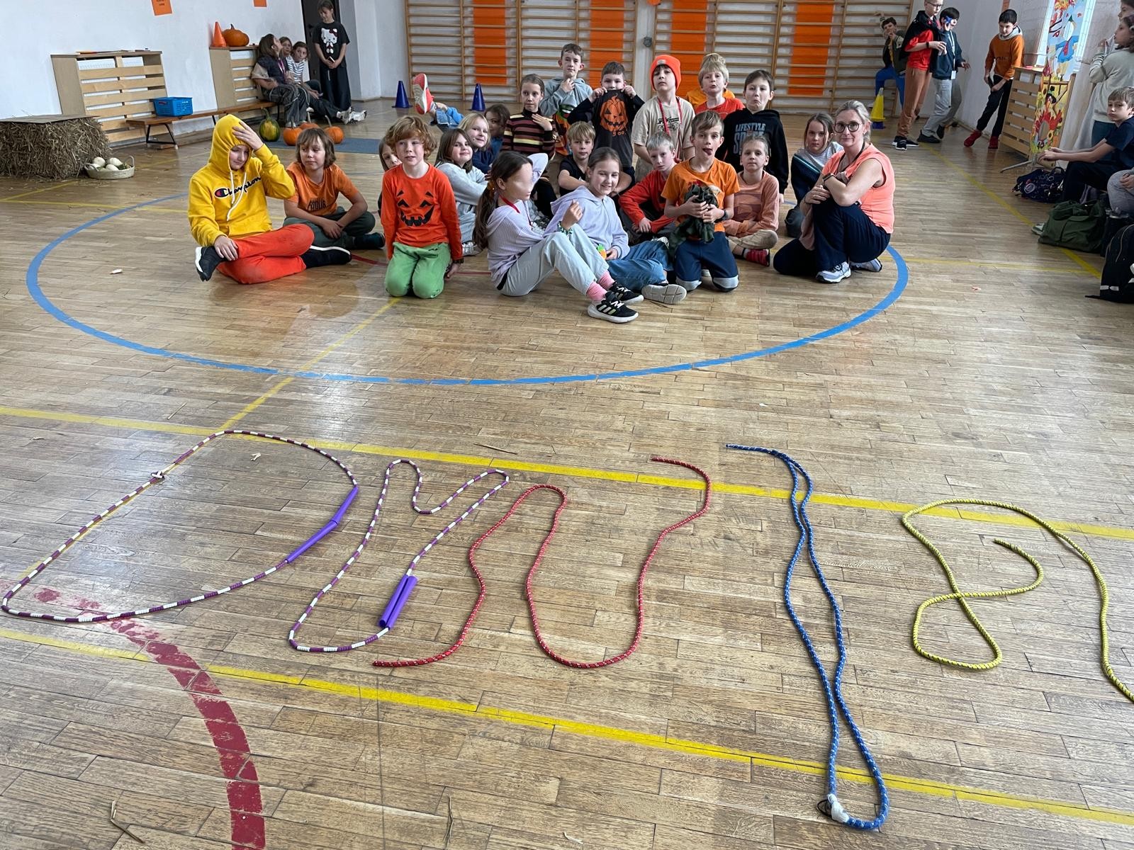 Grupa dzieci pozuje w sali gimnastycznej, wiele z nich ma pomarańczowe ubrania z motywami dyni. Na podłodze ułożono ze skakanek napis „DYNIA”.