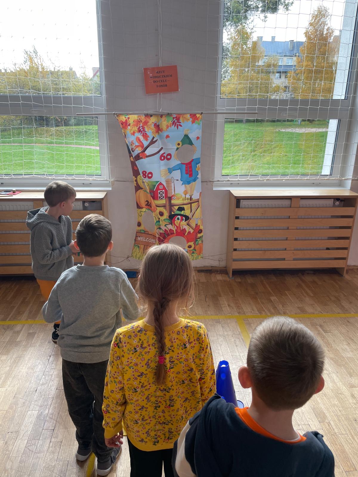 Czworo dzieci stoi tyłem, przygotowując się do rzutu do celu. Na wprost nich zawieszony jest kolorowy plakat z jesiennym motywem stracha na wróble i dyni, z punktowanymi polami („80”, „60”). Na ścianie wisi kartka z napisem informującym o grze: „RZUT WORECZKIEM DO CELU”.