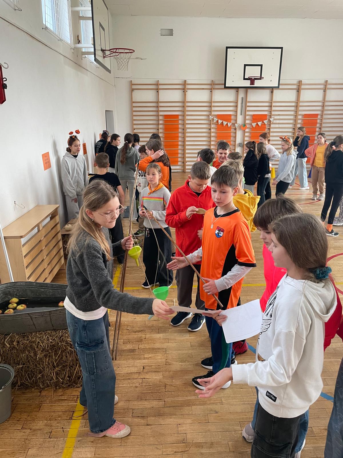 Kilkoro starszych dzieci uczestniczy w zadaniach. Dziewczynka na pierwszym planie w okularach i szarym swetrze podaje kijek innym dzieciom. W tle widać fragment stanowiska z sianem i skrzynią z jabłkami. 