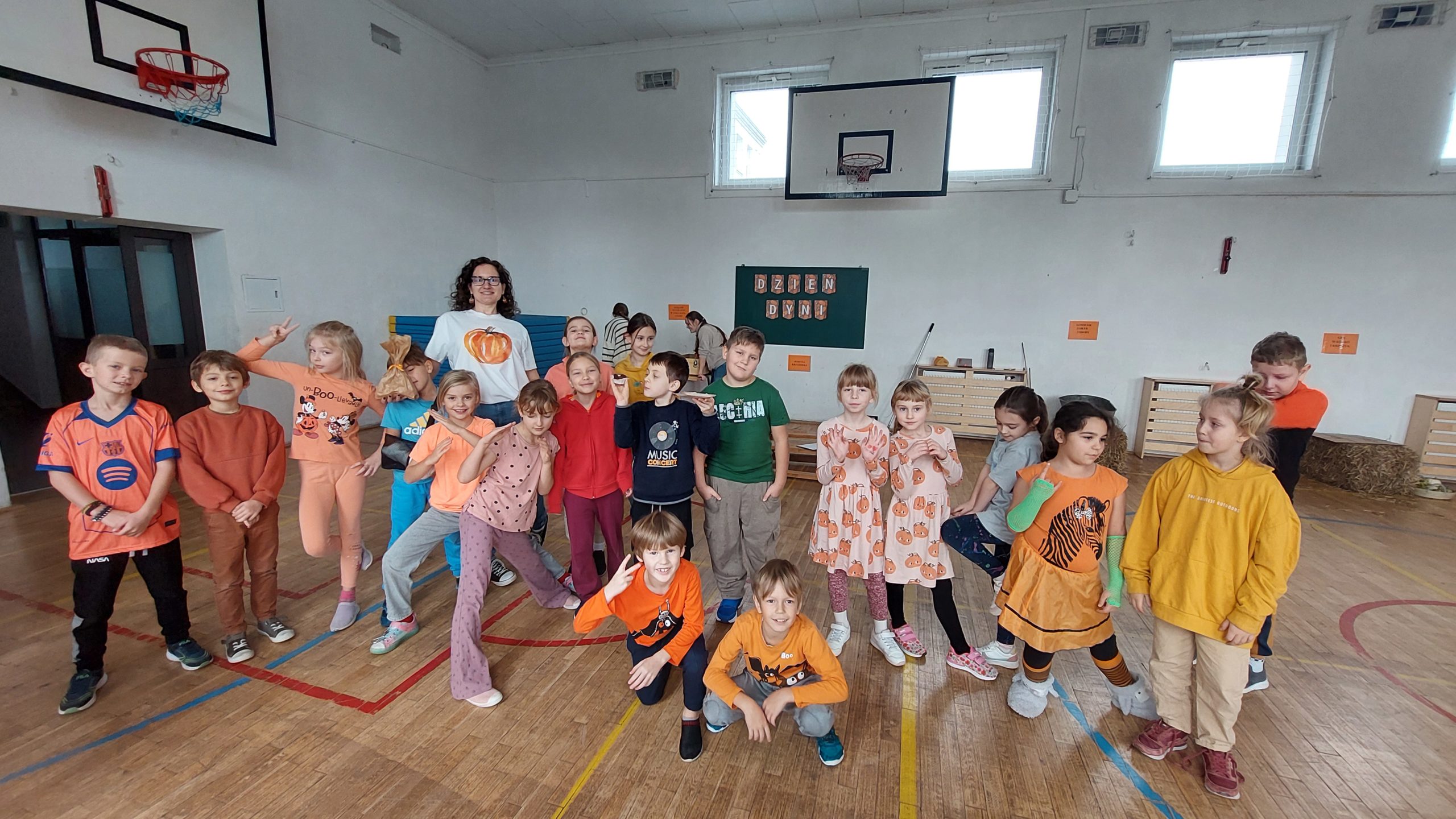 Duża grupa dzieci i jedna dorosła kobieta w białej koszulce z motywem dyni pozuje na sali gimnastycznej. Dzieci uśmiechają się i wykonują różne gesty.
