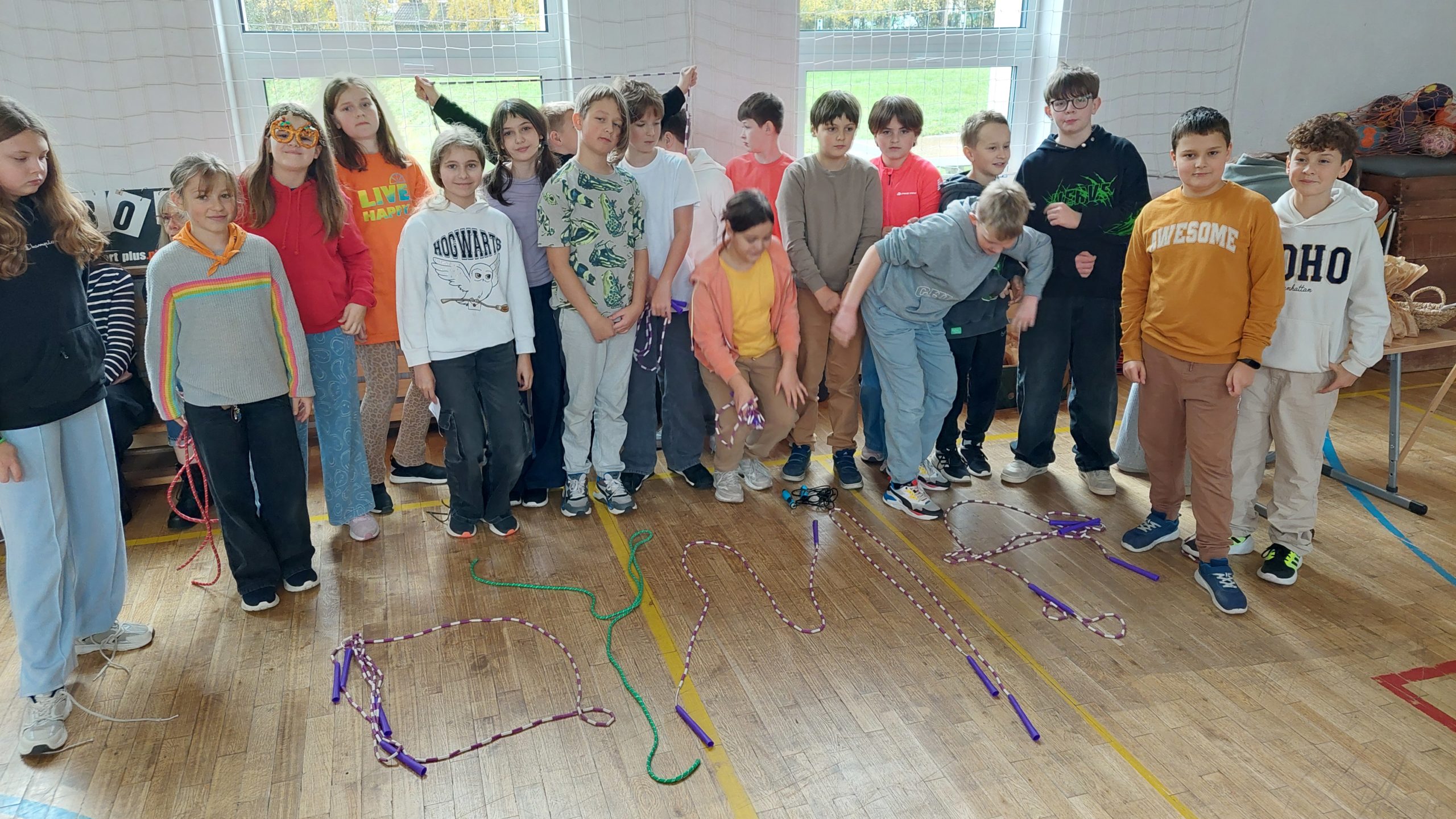 Grupa starszych dzieci i nastolatków pozuje w rzędzie na sali gimnastycznej. Na podłodze widać skakanki ułożone w napis „DYNIA”.