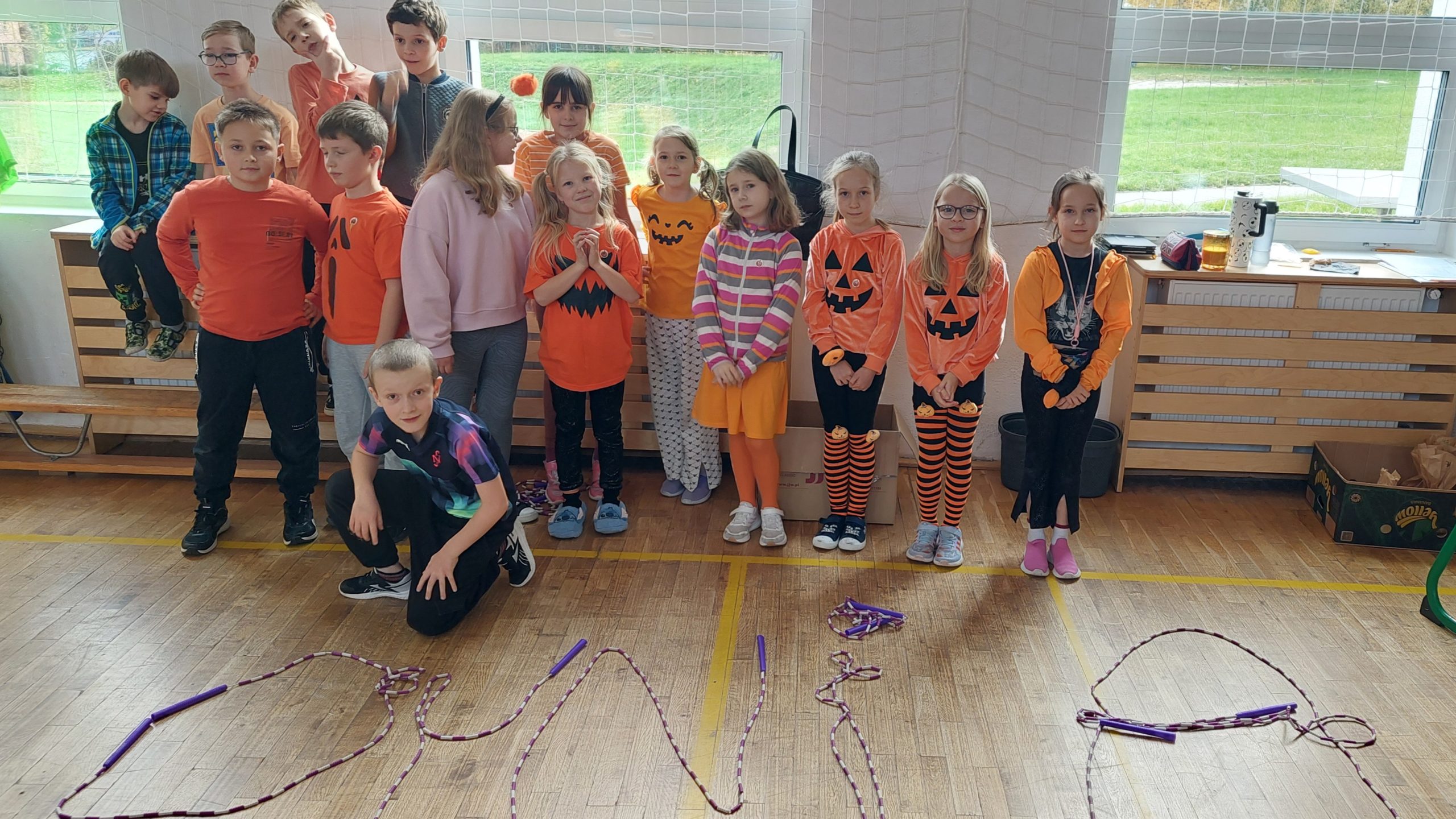 Grupa dzieci pozuje w sali gimnastycznej, wiele z nich ma pomarańczowe ubrania z motywami dyni. Na podłodze ułożono ze skakanek napis „DYNIA”.