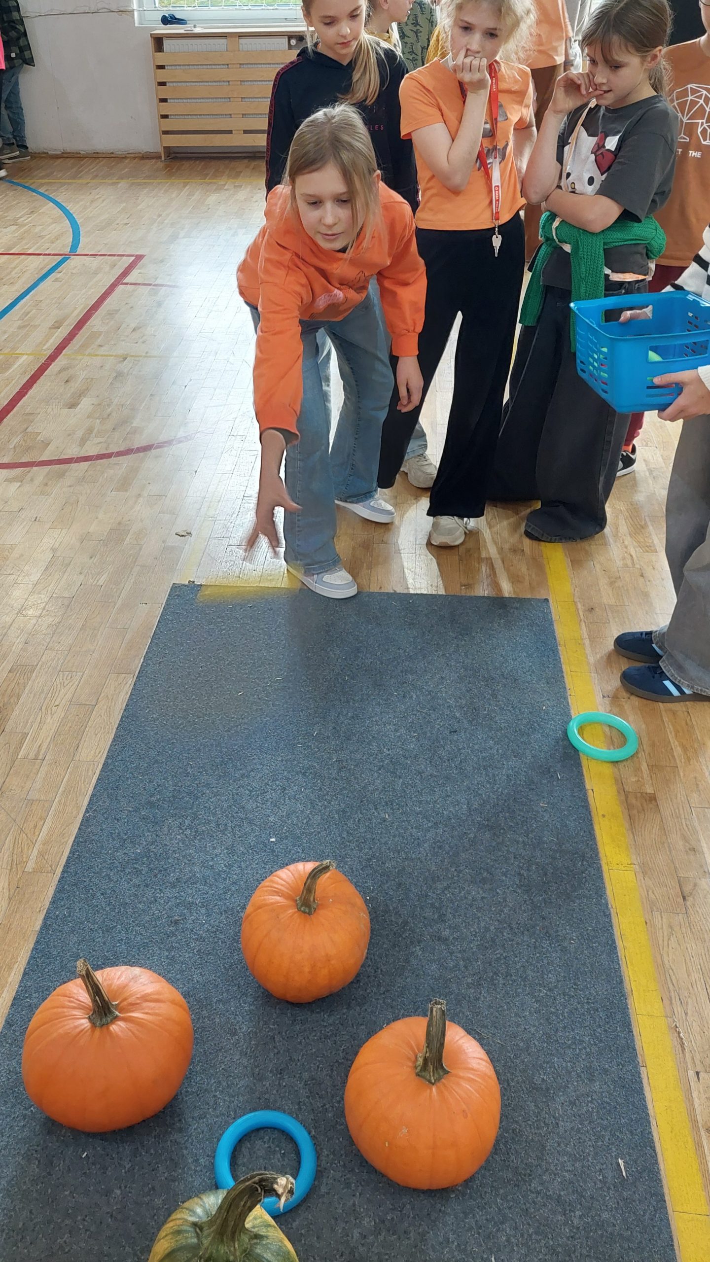 Dziewczynka w pomarańczowej bluzie rzuca obręcz w stronę kilku pomarańczowych dyń ustawionych na podłodze, a inne dzieci przyglądają się z boku.