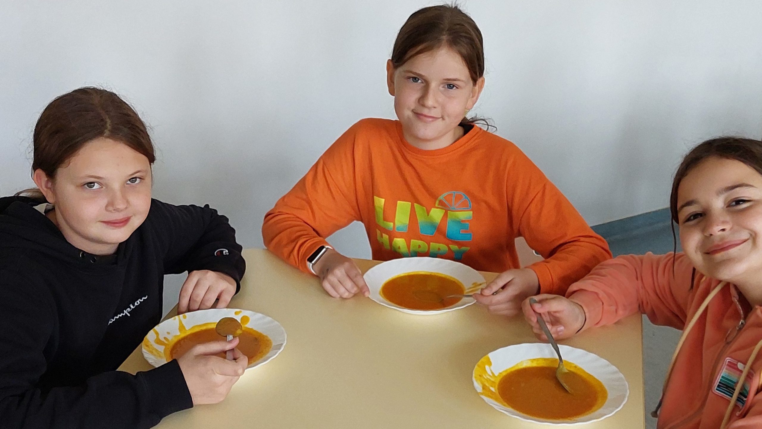 Trzy uśmiechnięte dziewczynki siedzą przy stole i jedzą zupę dyniową.