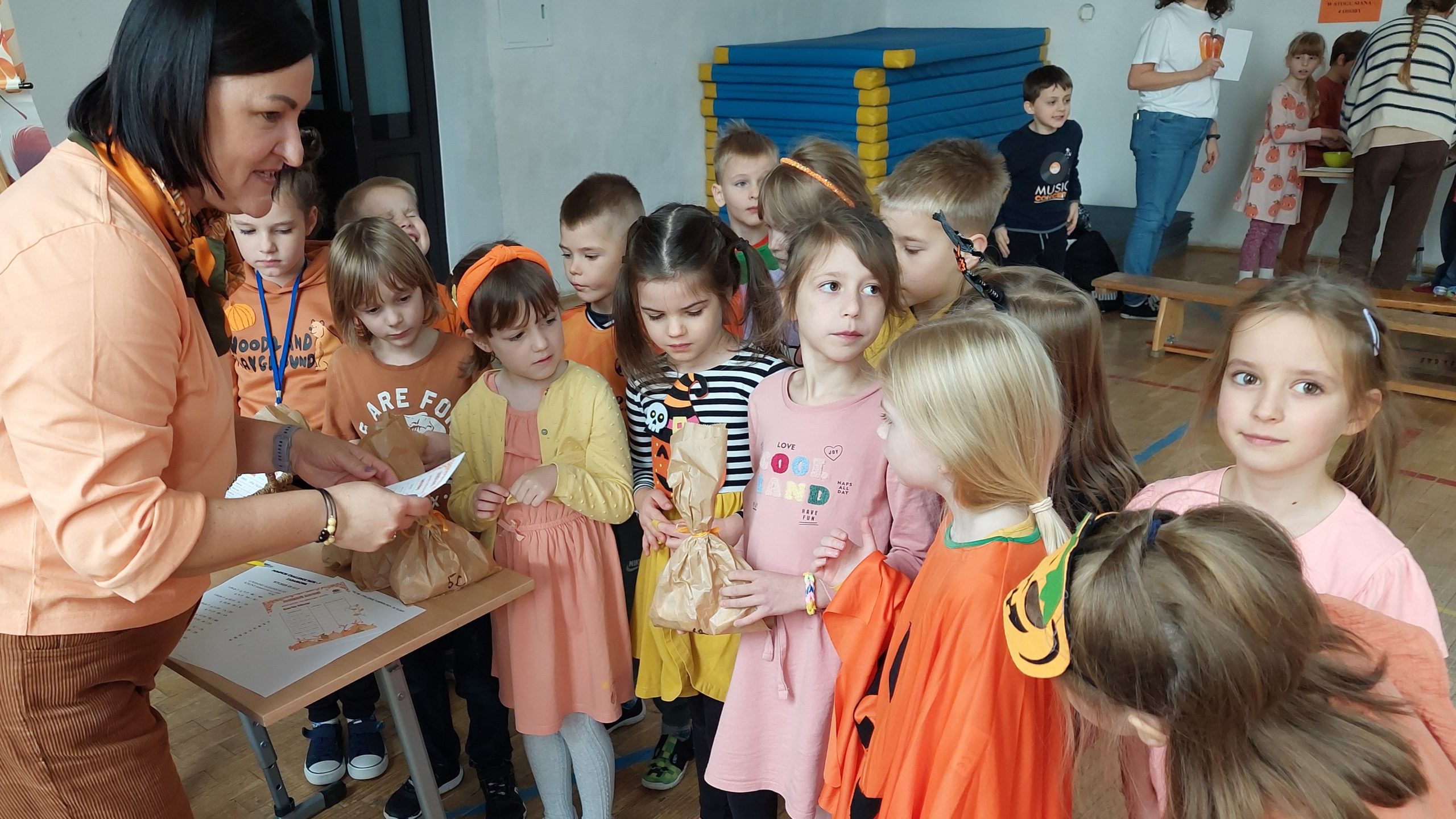Na sali gimnastycznej kobieta w pomarańczowej bluzce podaje małą papierową torebkę stojącej przed nią grupie małych dzieci. Dzieci są ubrane głównie w odcienie pomarańczowego, żółtego i różowego.