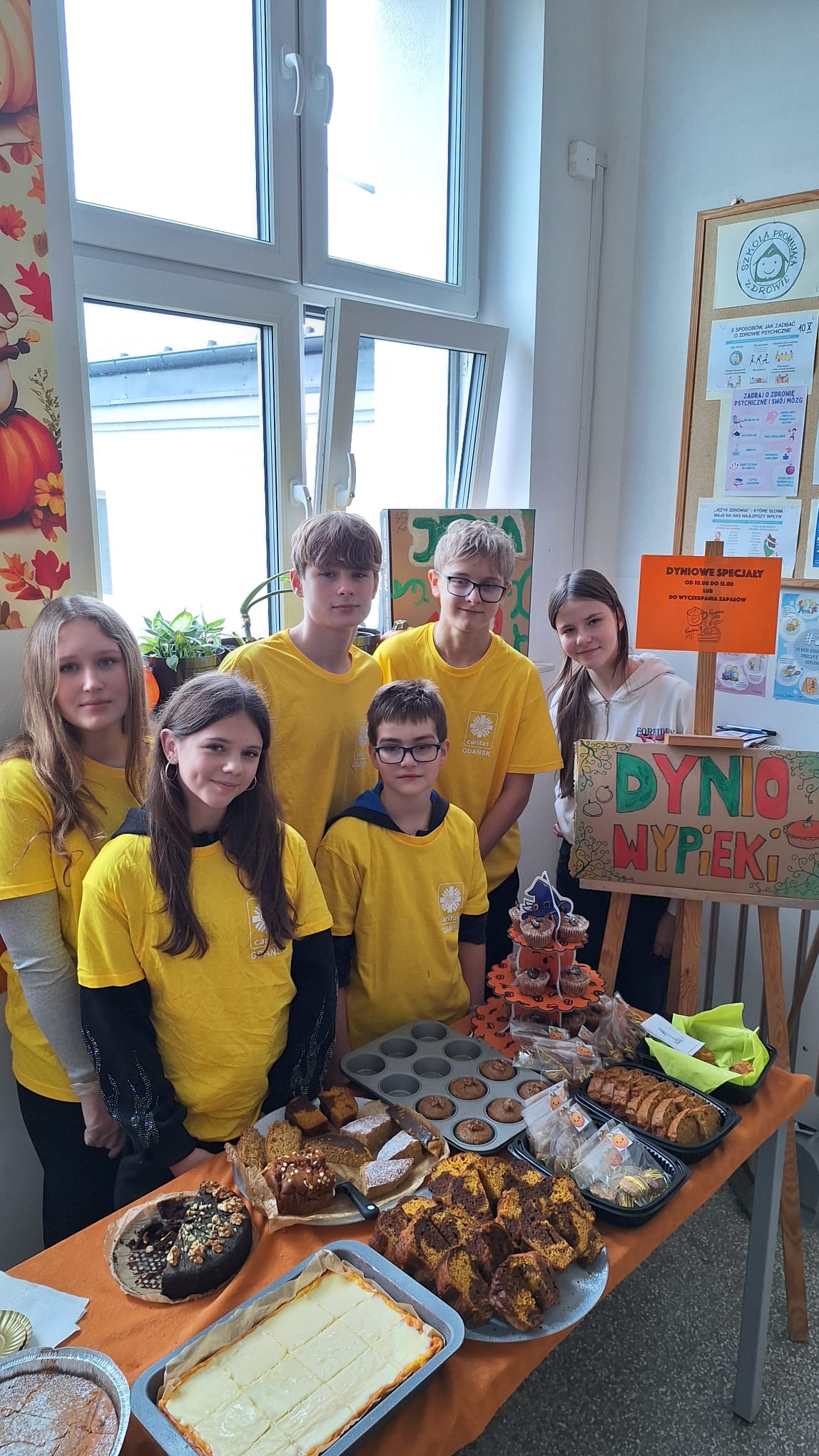 Pięciu nastolatków w żółtych koszulkach z logo stoi za stołem z dyniowymi wypiekami. Przed nimi widać duży wybór ciast i babek, a także babeczki. Jest to prawdopodobnie grupa wolontariuszy lub organizatorów.