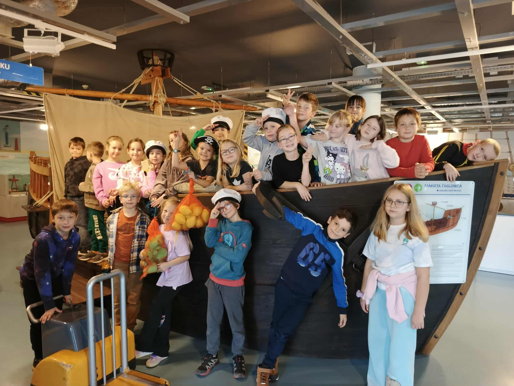 Grupa około dwudziestu kilku dzieci stoi i siedzi na dużej, drewnianej makiecie statku. Dzieci mają różne pozy — niektóre stoją na pokładzie, inne opierają się o burtę, a kilka siedzi z przodu statku. Niektóre dzieci mają czapki marynarskie lub pirackie, jedno dziecko trzyma worek z pluszowymi owocami i warzywami. W tle widać wnętrze sali muzealnej z wystawami o morzu i żegludze.