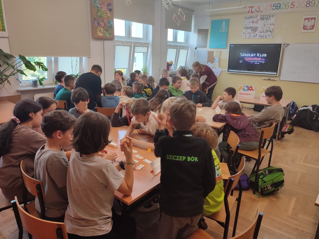 W sali lekcyjnej dzieci siedzą w grupach grając w grę Rummikub. W tle, na tablicy widnieje napis "Szkolny Klub Rummikub". Pod lampami wiszą bryły figur przestrzennych, na ścianach wiszą plakaty ze wzorami matematycznymi.