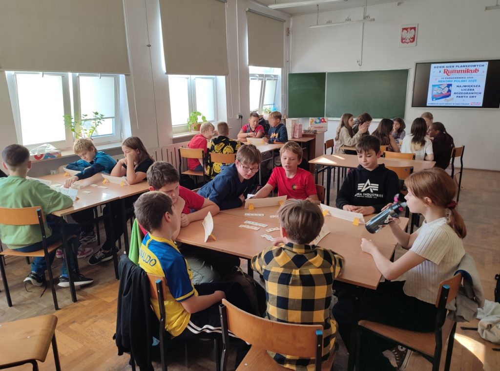 W sali lekcyjnej dzieci siedzą w grupach grając w grę Rummikub. W tle, na tablicy widnieje napis "Dzień Gier Planszowych z Rummikub. Największa liczba rozegranych partii gry".