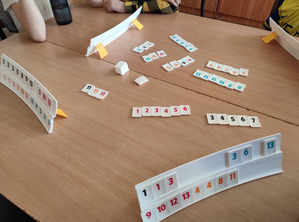 Zdjęcie przedstawia widok z góry na dwie złączone szkolne ławki, na których na środku rozłożone są grupy i serie liczb Rummikub.  Na zdjęciu są również cztery półeczki z kostkami graczy.