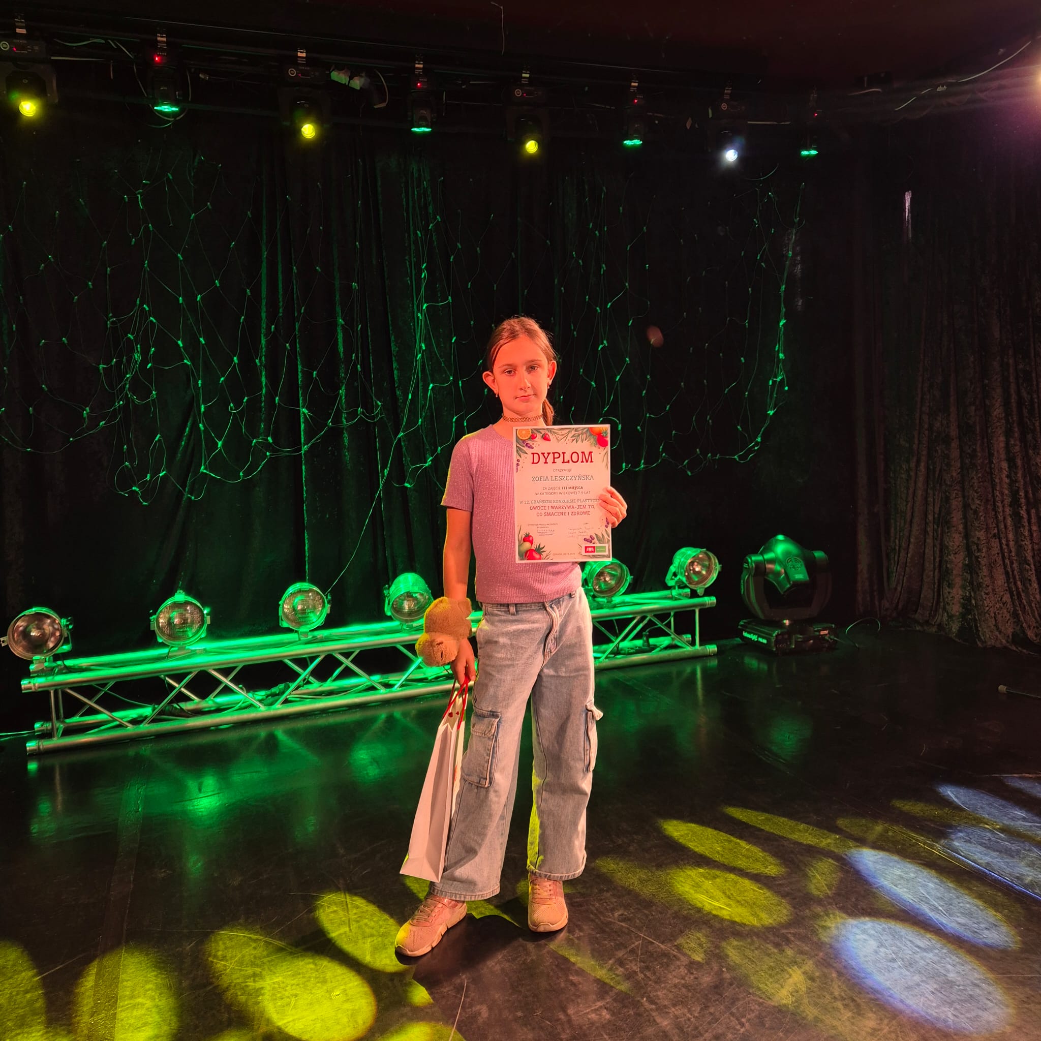 Uczennica z dyplomem w lewej ręce i torbą w prawej ręce stoi na scenie  na tle kolorowych lamp.
