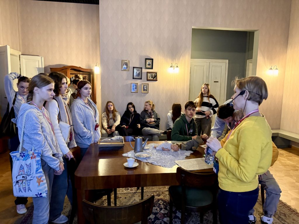 Grupa uczniów klasy 8b znajduje się w pomieszczeniu urządzonym w stylu dawnego mieszkania. Część z nich stoi wokół dużego drewnianego stołu, na którym leżą rekwizyty, w tym lalka i naczy-nia. Pozostali siedzą na kanapie i krzesłach pod ścianą. Przed grupą stoi przewodniczka w żółtym swetrze. Na ścianie wiszą małe rodzinne fotografie, a wnętrze oświetlają lampy.