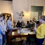 Grupa uczniów klasy 8b znajduje się w pomieszczeniu urządzonym w stylu dawnego mieszkania. Część z nich stoi wokół dużego drewnianego stołu, na którym leżą rekwizyty, w tym lalka i naczy-nia. Pozostali siedzą na kanapie i krzesłach pod ścianą. Przed grupą stoi przewodniczka w żółtym swetrze. Na ścianie wiszą małe rodzinne fotografie, a wnętrze oświetlają lampy.