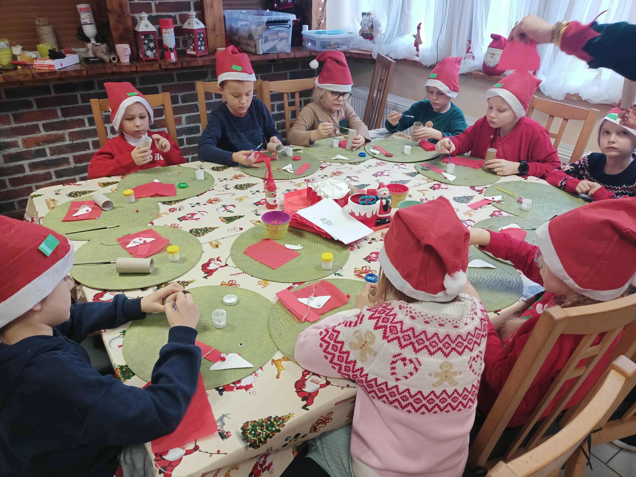 : Grupa dzieci w czerwonych czapkach Mikołaja siedzi przy dużym, świątecznie udekorowanym stole. Dzieci wykonują prace plastyczne, używając rolek po papierze, kolorowego papieru i kleju. Na stole znajdują się zielone podkładki, czerwone serwetki i różne materiały do prac ręcznych. W tle widać półki z dekoracjami świątecznymi.